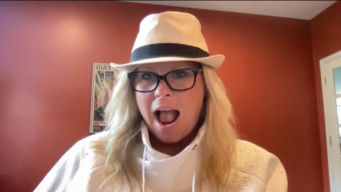 Amanda Heberg is a light skinned female with blonde wavy hair wearing a white fedora with a black stripe, black glasses wearing a white sweater