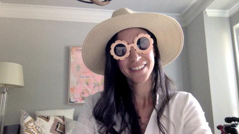 Laura Chaparro a light skinned female with long brown hair with a wide brimmed sun hat and fun sunglasses wearing a white shirt smiling at the camera