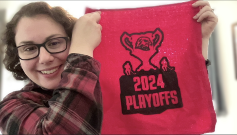 Madeline Jensen is a light skinned female with short curly brown hair, brown eyes, glasses wearing a red and black flannel shirt smiling at the camera holding a 2024 hockey playoffs towel