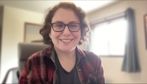 Madeline Jensen is a light skinned female with short curly brown hair, brown eyes, glasses wearing a red and black flannel shirt smiling at the camera