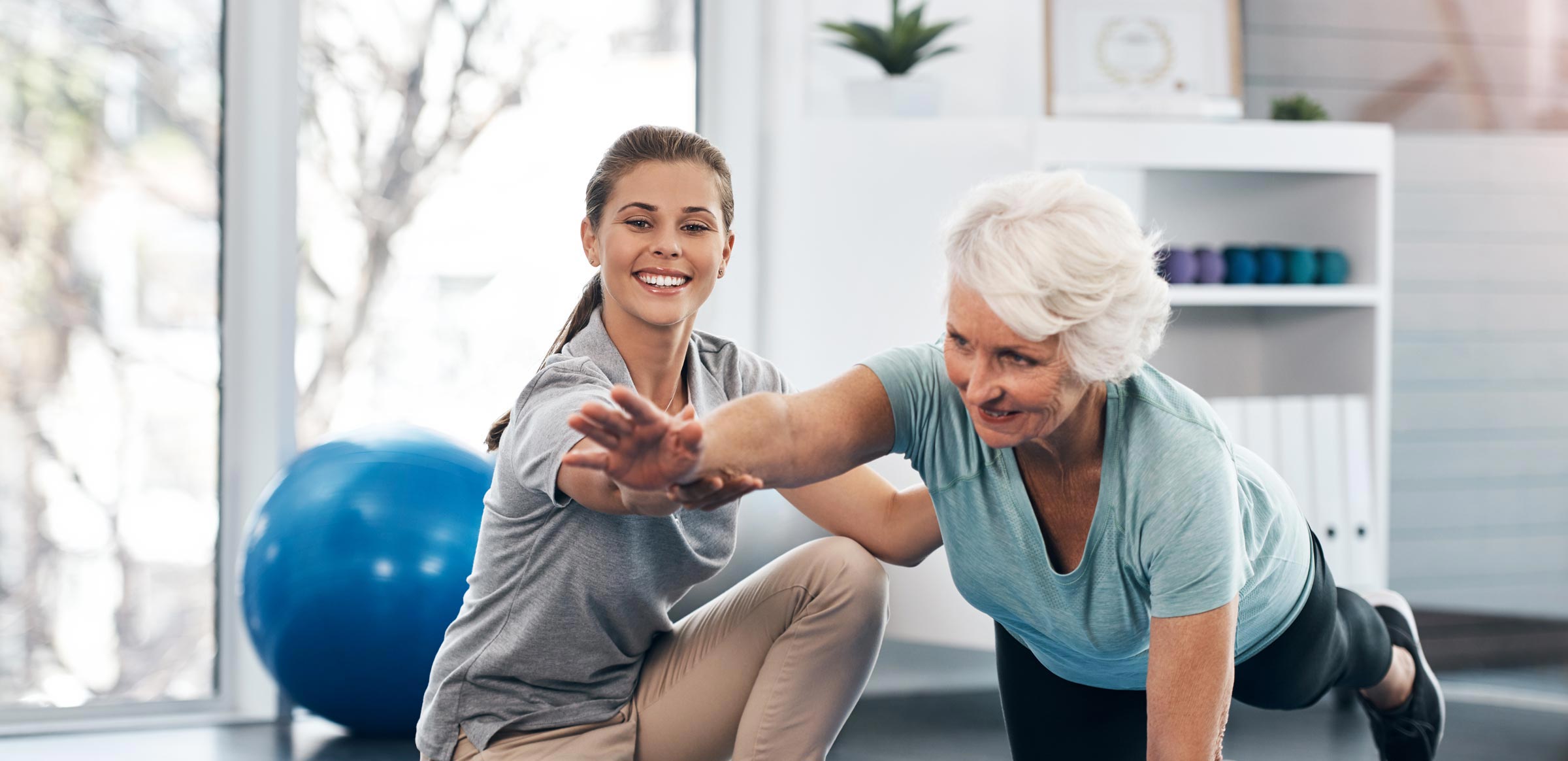 A light-skinned, female presenting therapist assisting a white-haired light-skinned female presenting patient with a floor exercise on a blue yoga mat