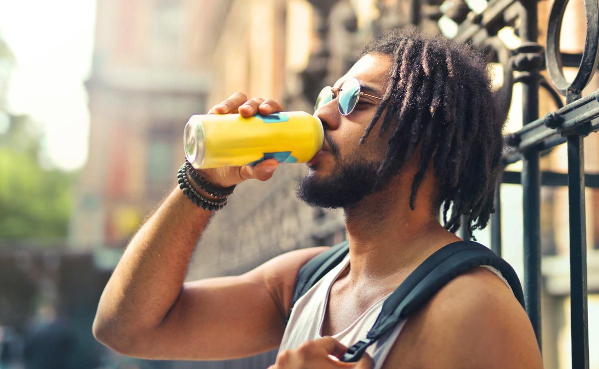A dark-skinned, male-presenting person on a residential street with sunglasses drinking out of a large yellow can