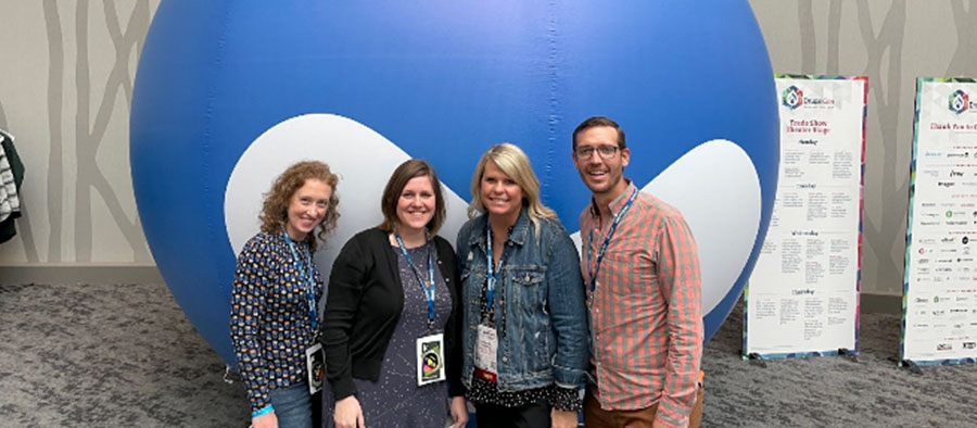 Three female-presenting people and one male-presenting person in front of a large blue inflatable droplet, all smiling at the camera.