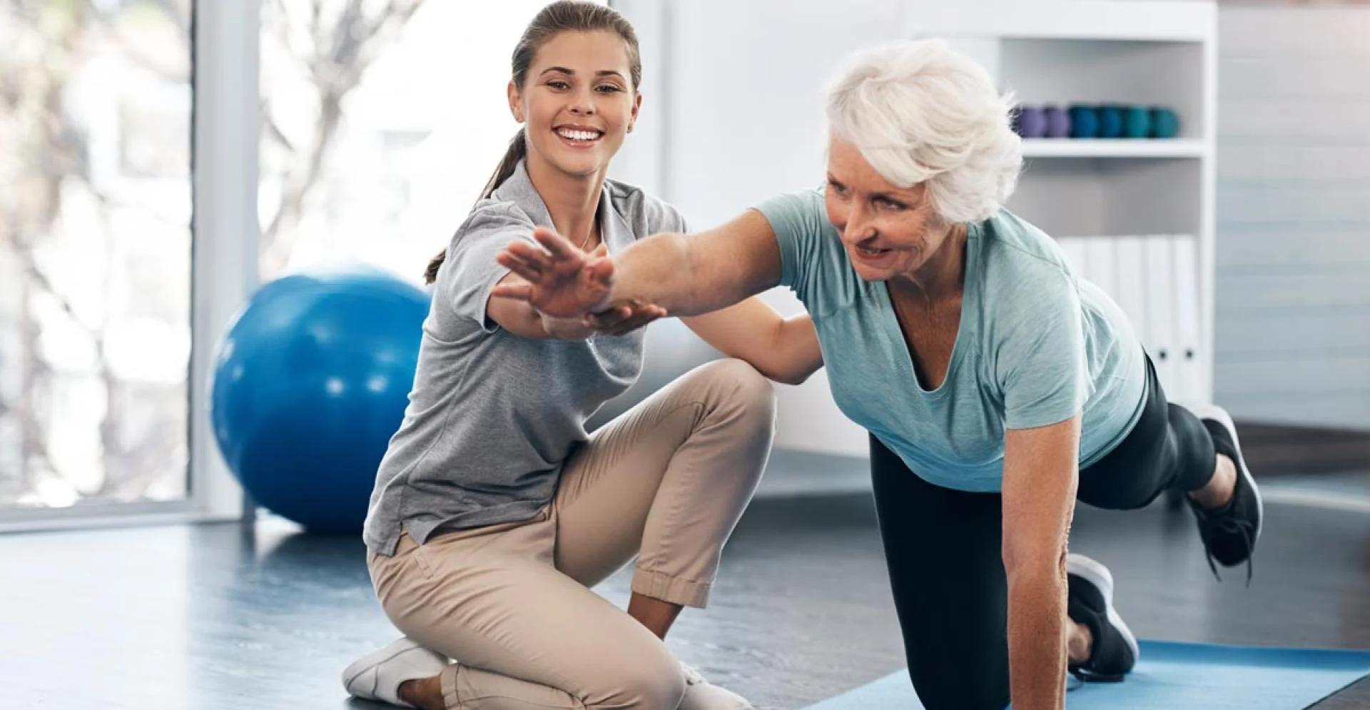 A light-skinned, female presenting therapist assisting a white-haired light-skinned female presenting patient with a floor exercise on a blue yoga mat