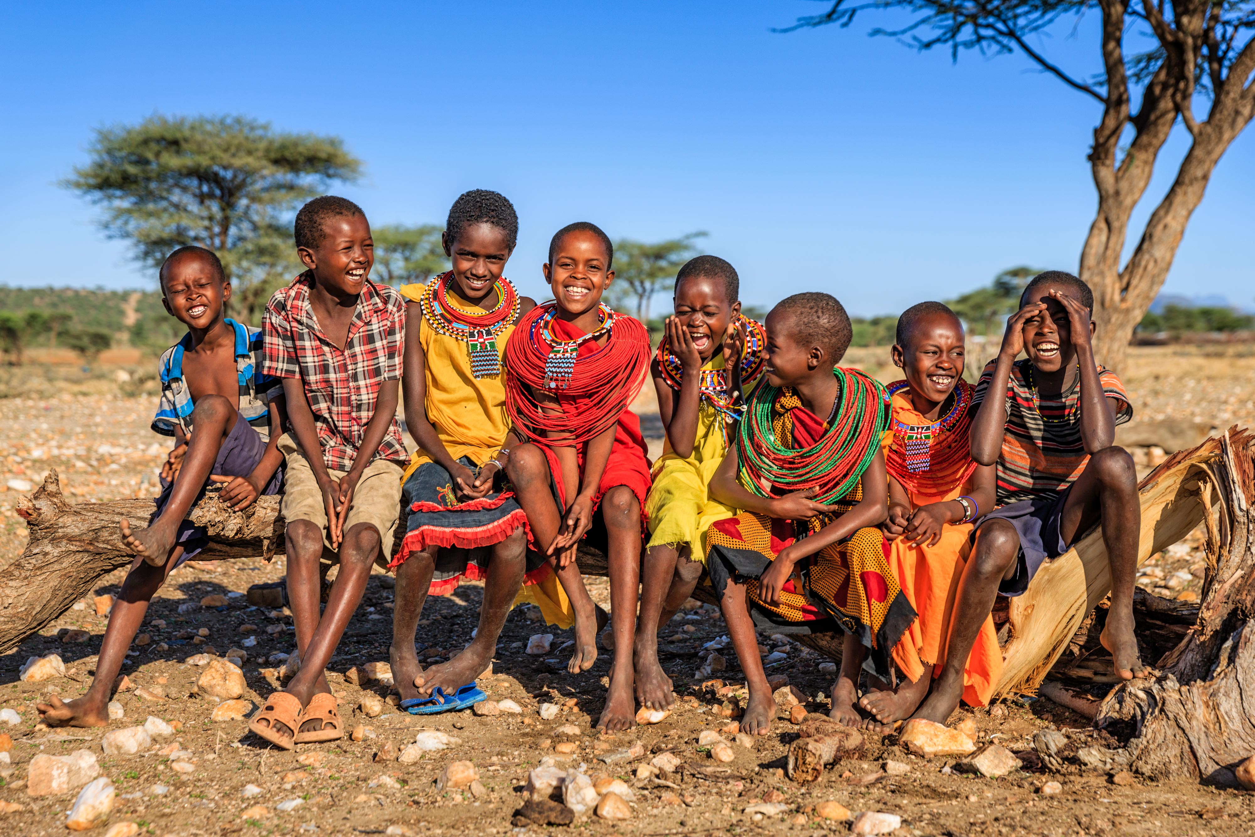 A group of eight African children sitting on a log in colorful clothing.