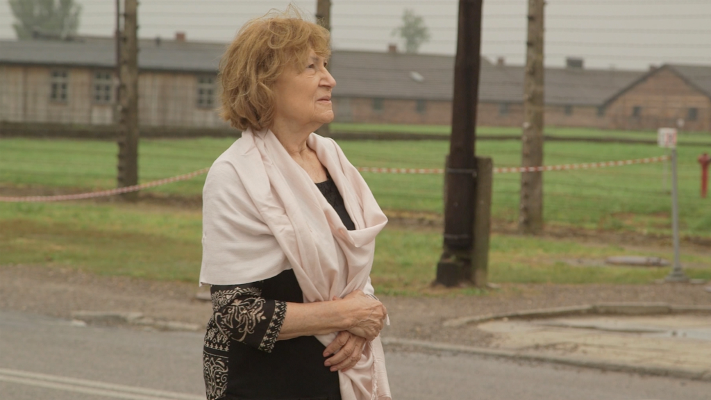 Woman with a shawl stands outdoors, looking to the side, with historic buildings behind.