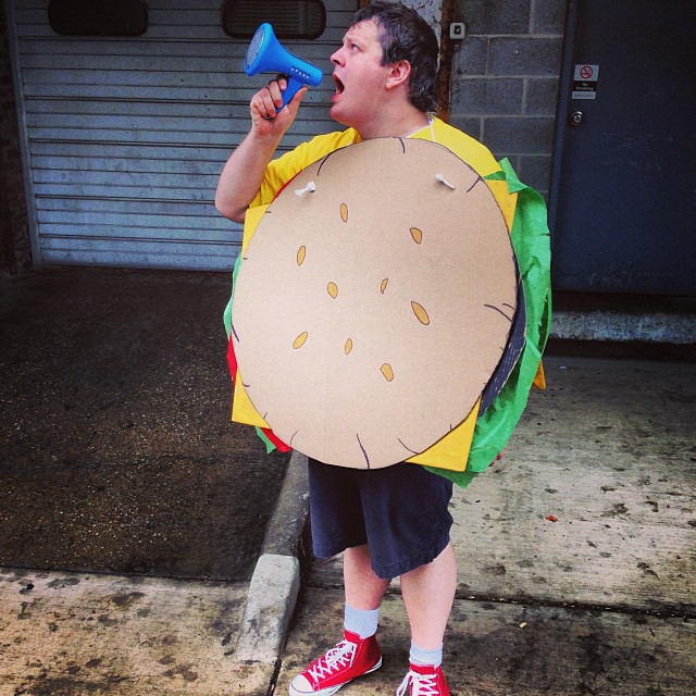 A male-presenting person in a hamburger costume shouting into a bullhorn outdoors.