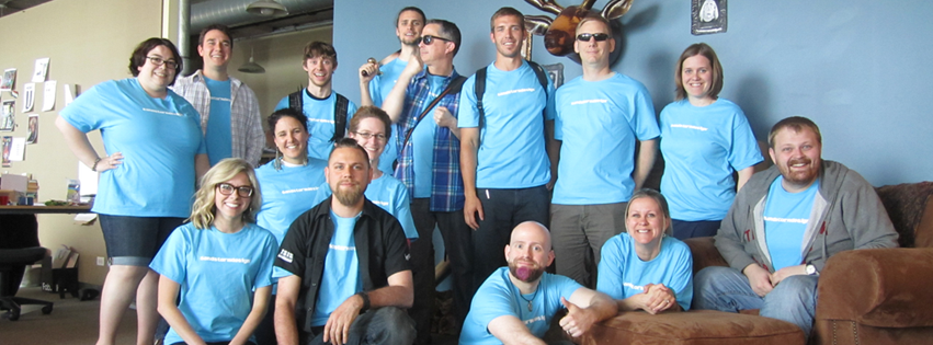 A group of thirteen people, wearing matching blue shirts, posing together as a team indoors. They are standing and sitting, smiling at the camera