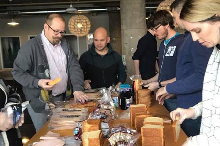 A group of people stand around a desk in an office making sandwiches.