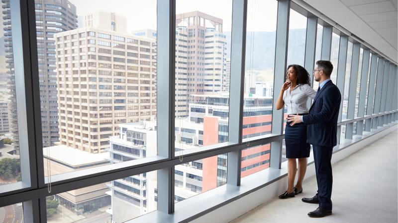One female and one male real estate professional standing in an empty office looking out windows at a city scape.