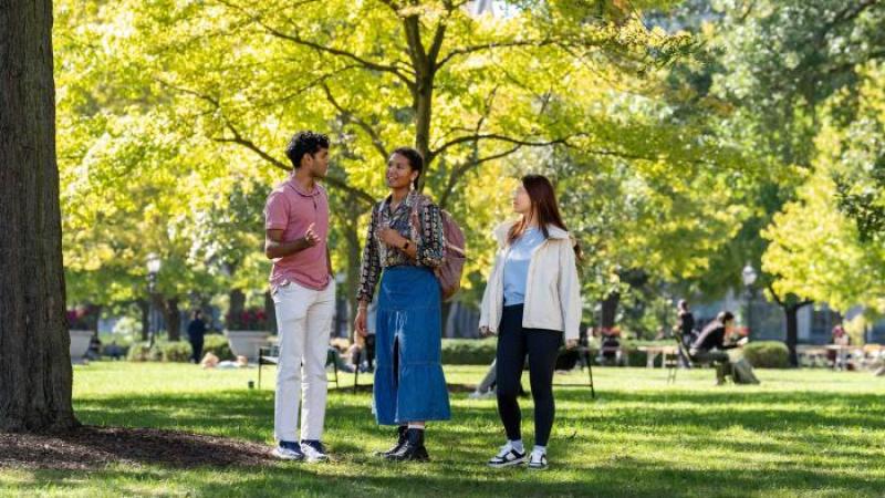 Three students talking and smiling in a sunny campus setting.