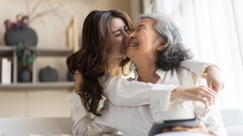 Woman hugs and kisses older woman, both smiling warmly in a cozy setting.