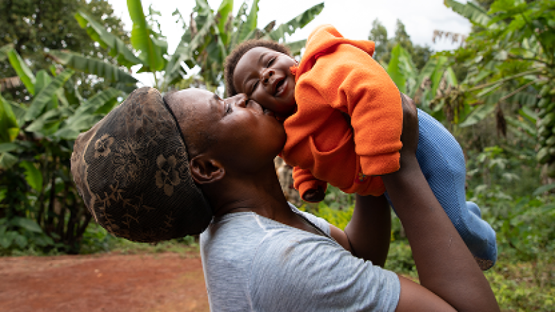 Mother holding and kissing her smiling baby in a lush, green setting.