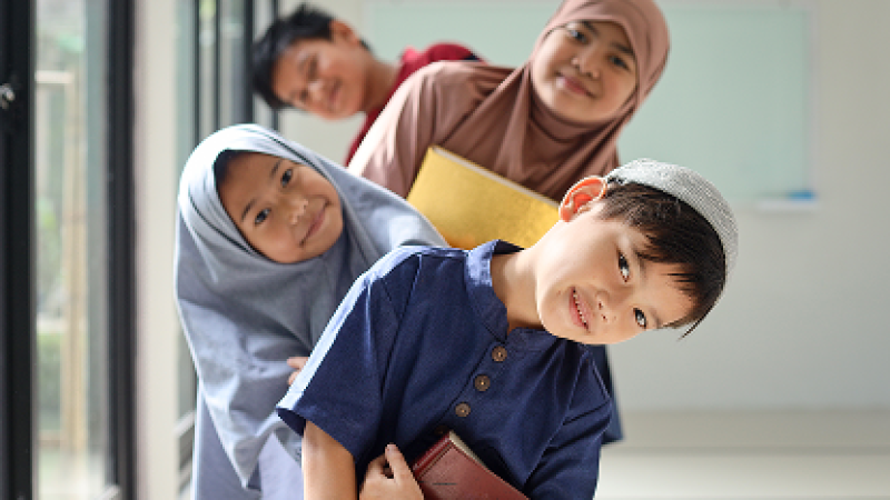 Kids smiling, leaning sideways in a line indoors, holding books.
