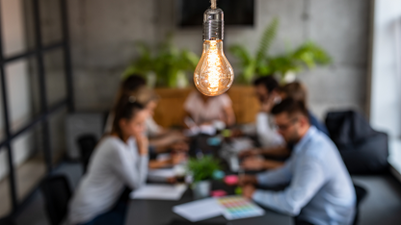 Close-up of light bulb with blurred group of people working at a table in the background.