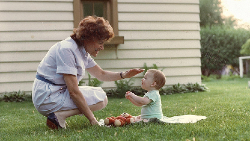 Archival photograph of Fritzie Fritzshall crouching on a lawn, smiling and reaching toward a seated baby during playtime.