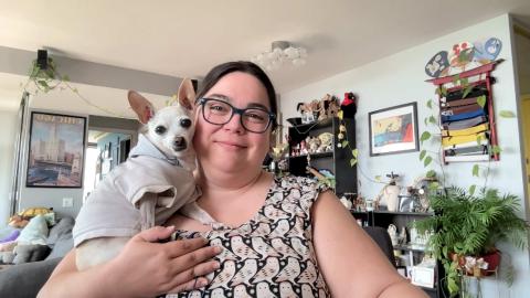 Alma Meshes a light skinned female with dark hair, blue glasses, wearing an owl print dress with plants, knick knacks and art work in the background holding her chihuahua, Chuppie