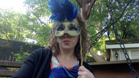 Anne Lentino is a light skinned female with curly red hair holding a masquerade mask in front of her face