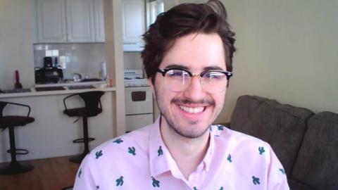 Cameron Panice a light skinned male with glasses, dark brown hair, dark brown mustache and stubble wearing a pink shirt with cactuses and a kitchen and couch in the background