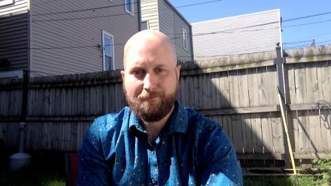Eric Savage is a light skinned male with blonde mustache and beard, bald head wearing a blue shirt with flowers with a fence and grass in the background