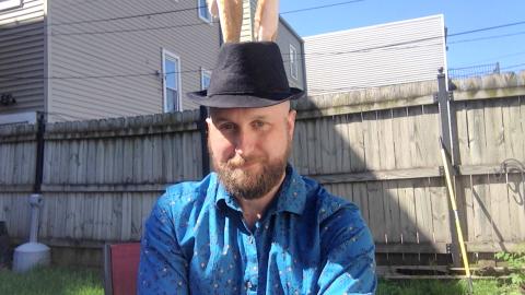 Eric Savage is a light skinned male with blonde mustache and beard, with a fedora with bunny ears on his head, wearing a blue shirt with flowers with a fence and grass in the background