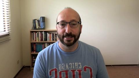 Jay Silverman is a light skinned male with dark mustache and beard, bald head, wearing glasses and a blue Chicago Cubs shirt with a book case in the background