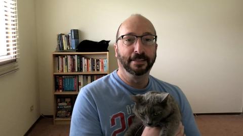 Jay Silverman is a light skinned male with dark mustache and beard, bald head, wearing glasses and a blue Chicago Cubs shirt holding his grey cat with a book case in the background with his black cat on top