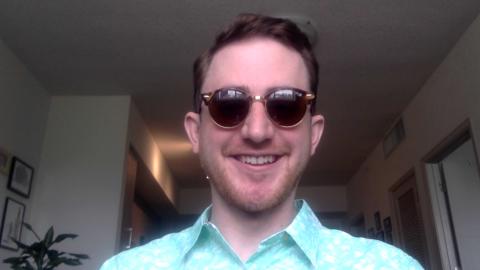 Neil Pichan is a light skinned male with brown hair, light brown facial hair, sun glasses, wearing a light green shirt with white flowers with a hallway in the background