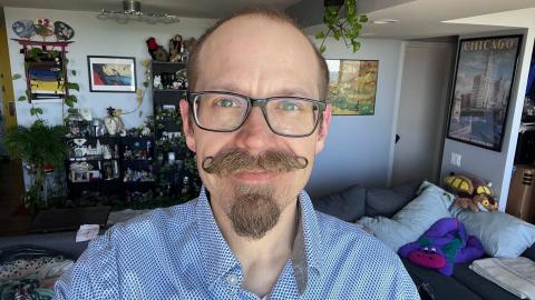 Nick Meshes a light skinned mail with black glasses, blonde hair, blonde handle bar mustache and goatee wearing a white shirt with blue diamonds with a book case of collectables and art work in the background