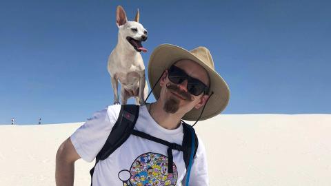 Nick Meshes a light skinned mail with sunglasses, tan colored hat, blonde handle bar mustache and goatee wearing a t-shirt holding faun colored chihuahua, Chuppie on his left shoulder in White Sands New Mexico