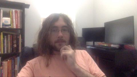 Noah Sekreter a light skinned male with glasses, light brown goatee and beard, long wavy light brown hair wearing a pink shirt with book case and computer monitors in the background