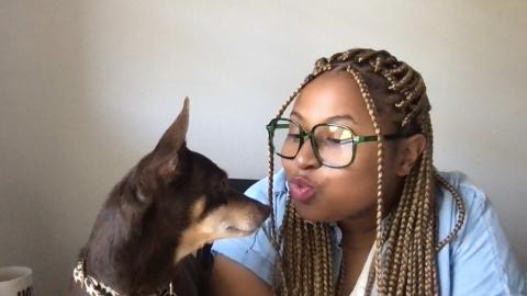 Sandie Starr is a dark skinned female with blond and brown braids with glasses wearing a white shirt with a jean short sleeve jacket over top air kissing her brown small dog, Gouda