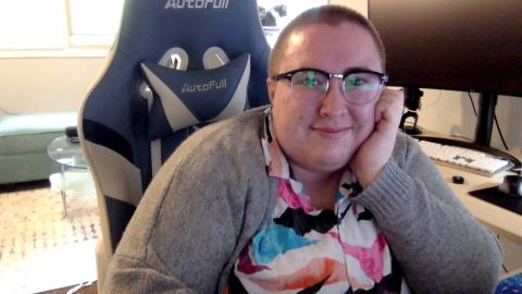 Syd Hunsinger is a light skinned person with glasses and a shaved head wearing a grey sweater and colorful shirt sitting in a gaming chair with a monitor in the background
