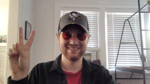 David Boocock a light skinned male wearing a penguins pride baseball cap, red colored sunglasses making a peace sign with his hand