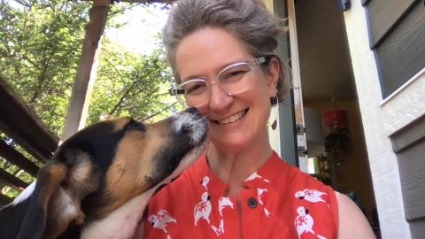 Janna Fiester a light skinned female with grey hair in an updo, clear glasses wearing a red shirt with birds on it smiling at the camera with her beagle, Miles