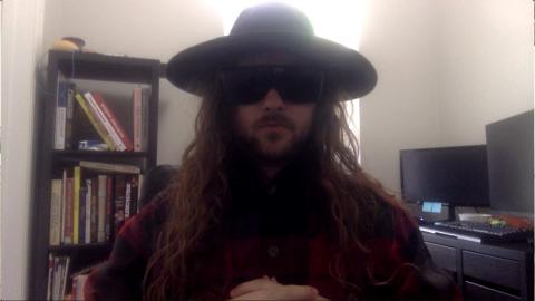 Noah Sekreter a light skinned male with large dark sunglasses, light brown goatee and beard, long wavy light brown hair with a black wide brimmed hat wearing a large brimmed black hat with book case and computer monitors in the background