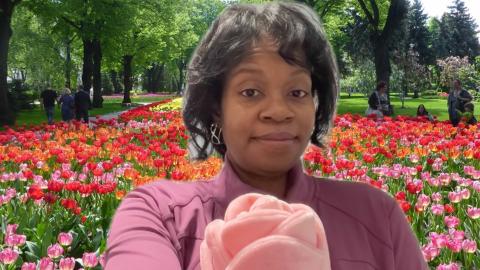 Maria Kelly is a dark skinned female with shoulder length black hair, wearing earrings and a purple shirt handing a tulip to the viewer in a field of tulips