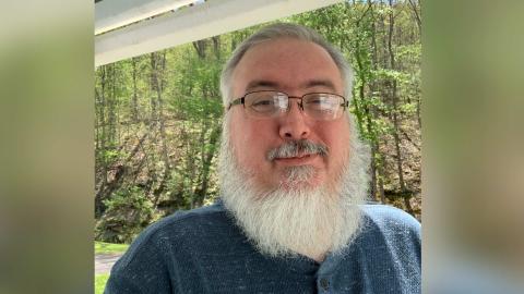 Shawn Hopkins a light skinned mail with glasses, a white beard and mustache, wearing a blue shirt, and smiling at the camera with a wooded area behind him