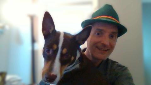 John Brooks a light skinned male with brown hair smiling at the camera wearing a green hat with his brown dog with a white stripe on his face sitting in his lap