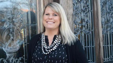 Amanda Heberg is a light skinned female with blonde hair wearing a black shirt and sweater, silver multi layer necklace standing in front of a decorative glass door