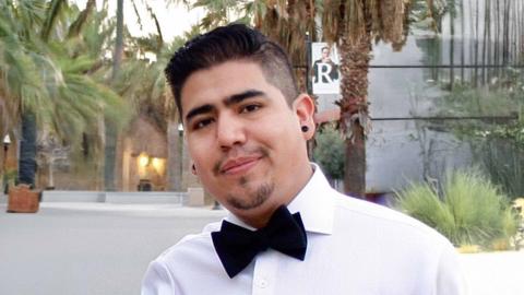 Seb Rodriguez is a male with dark spiky hair, goatee wearing a white dress shirt with a black bowtie standing in front of palm trees