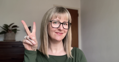 Amber Reeves is a light skinned female with shoulder length blonde hair, black glasses, wearing a green shirt smiling at the camera making a peace sign