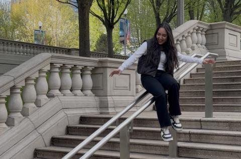 Isha is sliding down a stair railing, trees in the background.