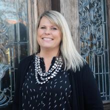 Amanda Heberg is a light skinned female with blonde hair wearing a black shirt and sweater, silver multi layer necklace standing in front of a decorative glass door