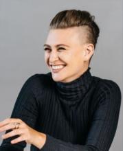 Devin Owsley-Aquilia: light-skinned non-binary person smiling, with dark blonde hair pulled back, wearing a black turtleneck against a grey wall