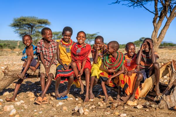 Group of 8 smiling children from the Samburu tribe in Kenya sitting on a tree trunk wearing colorful clothing.