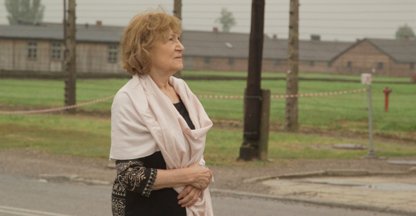 Elderly woman standing outside in front of historic buildings, wearing a light shawl.